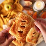 A hand holding up a golden-brown puff pastry apple hand pie with apples, a gold napkin, and a candle on a grey table.