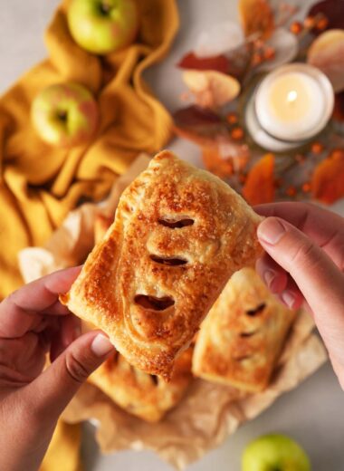 A hand holding up a golden-brown puff pastry apple hand pie with apples, a gold napkin, and a candle on a grey table.