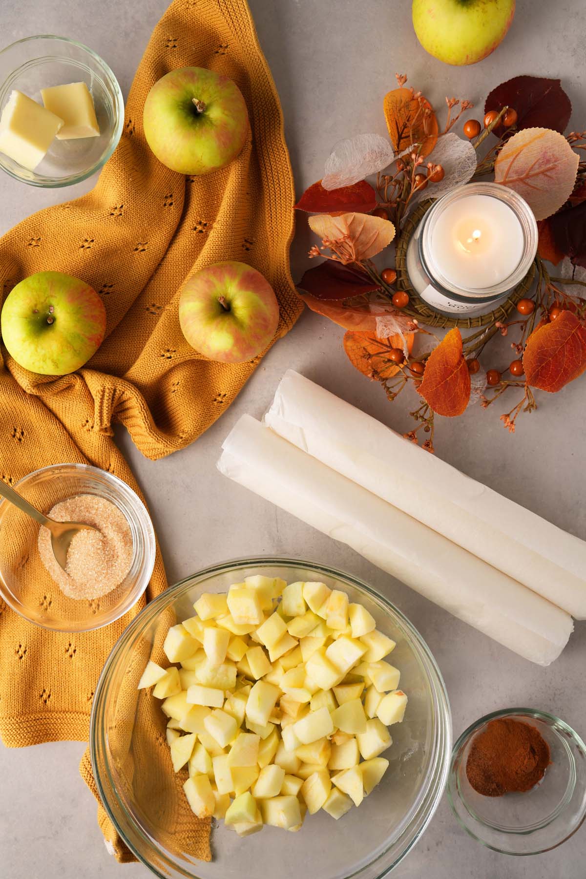 Apple hand pies ingredients on a grey table with apples, dough, and a candle near a fall candle ring.