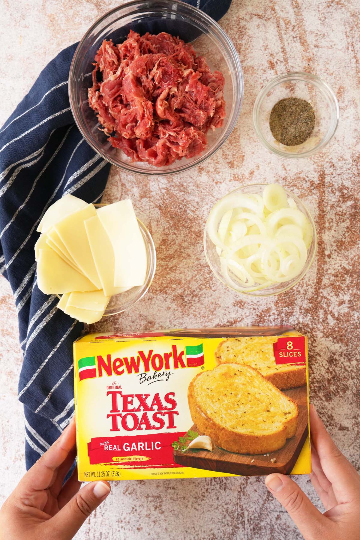 A hand holding a New York Bakery Texas Toast box near ingredients in glass bowls near a blue napkin.