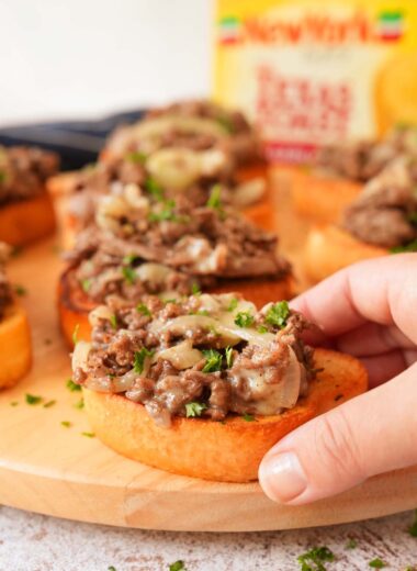 A hand holding a steak and cheese texas toast with parsley on top. A box of New York Bakery Toast is in the background.