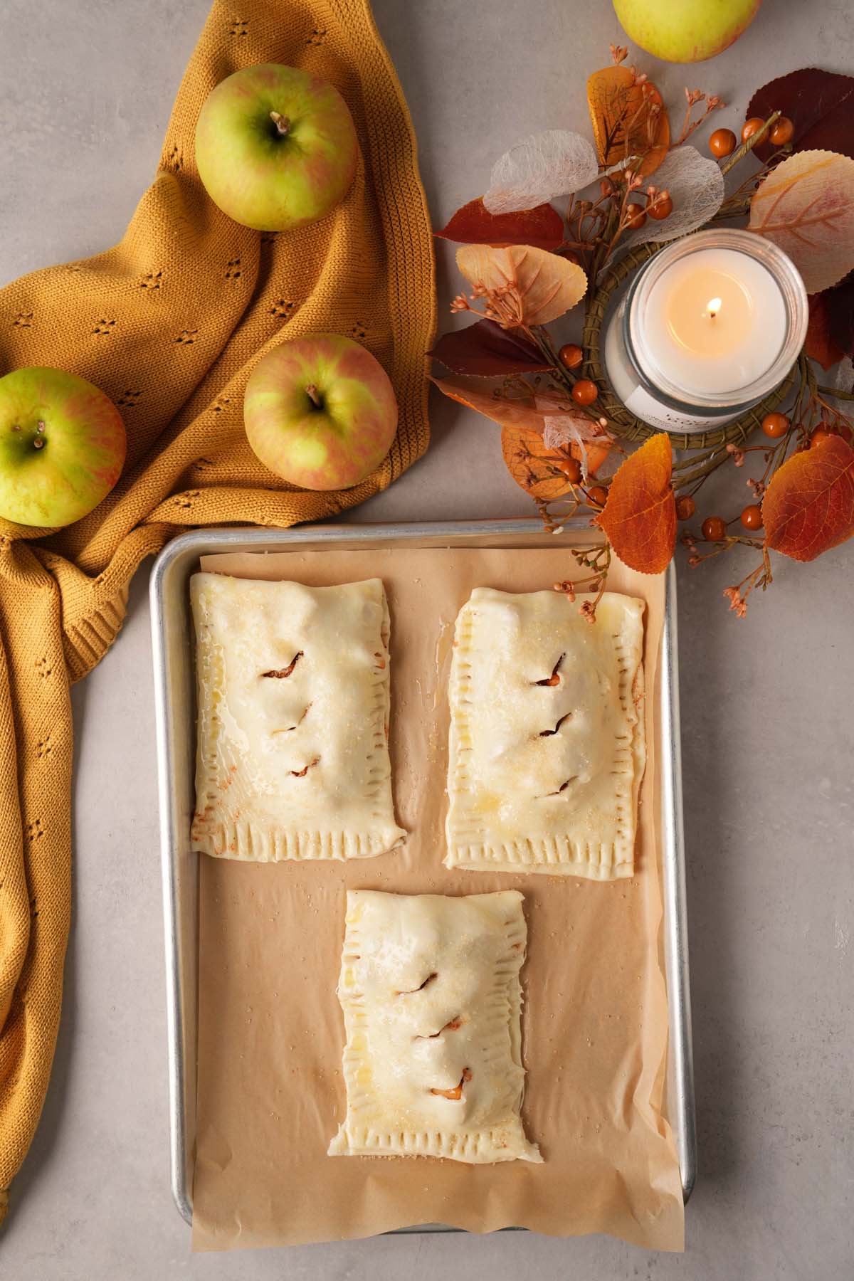 Three unbaked apple hand pies on a parchment-lined pan with apples, a gold napkin, and a fall candle in the background.
