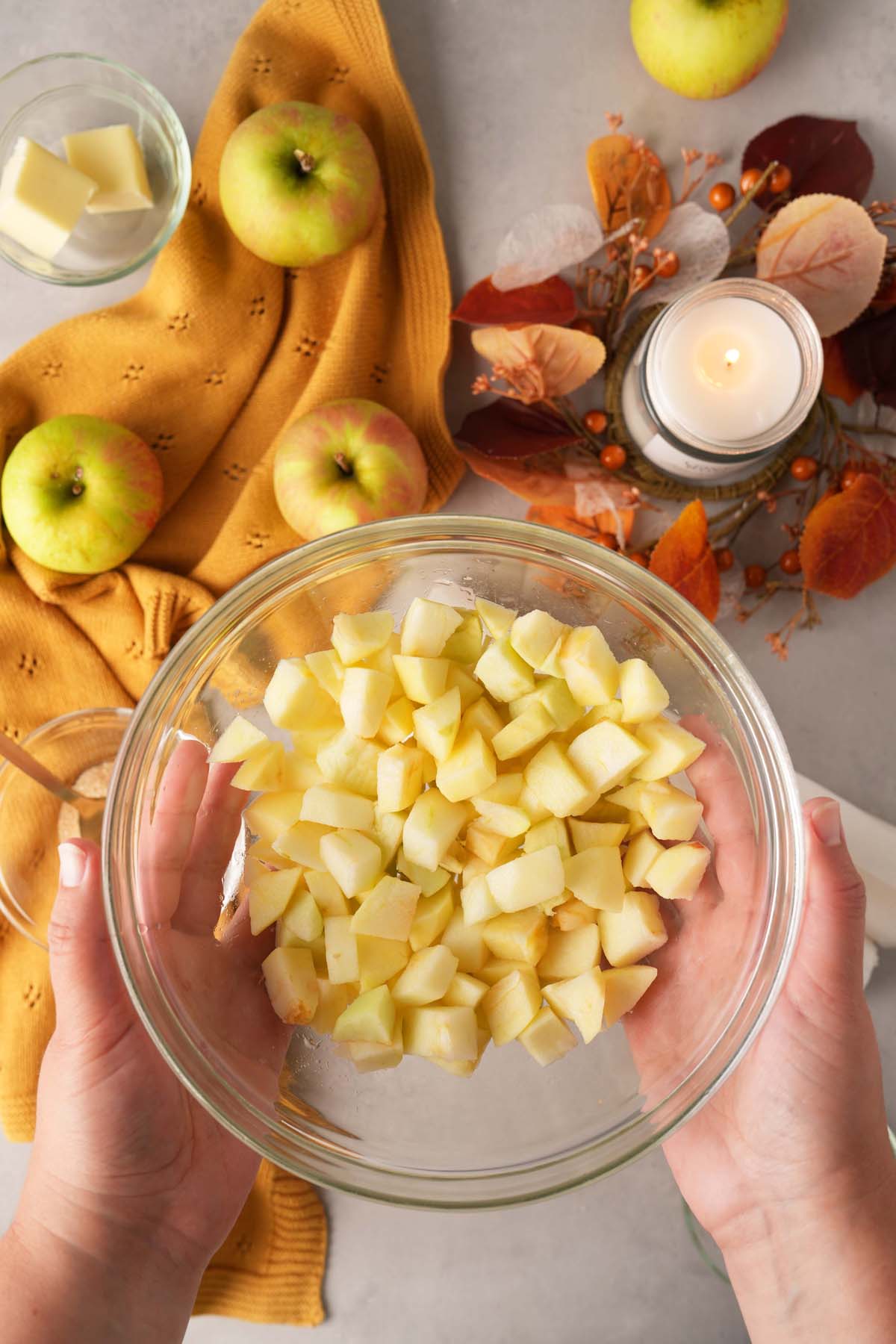 Hand holding up a glass bowl with diced apples. There are apples and a fall candle in the background.