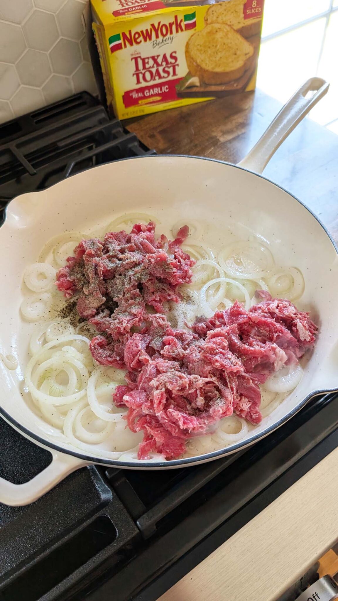 Seasoned, shaved steak in a skillet with onions. A box of New York Bakery Toast in the background.