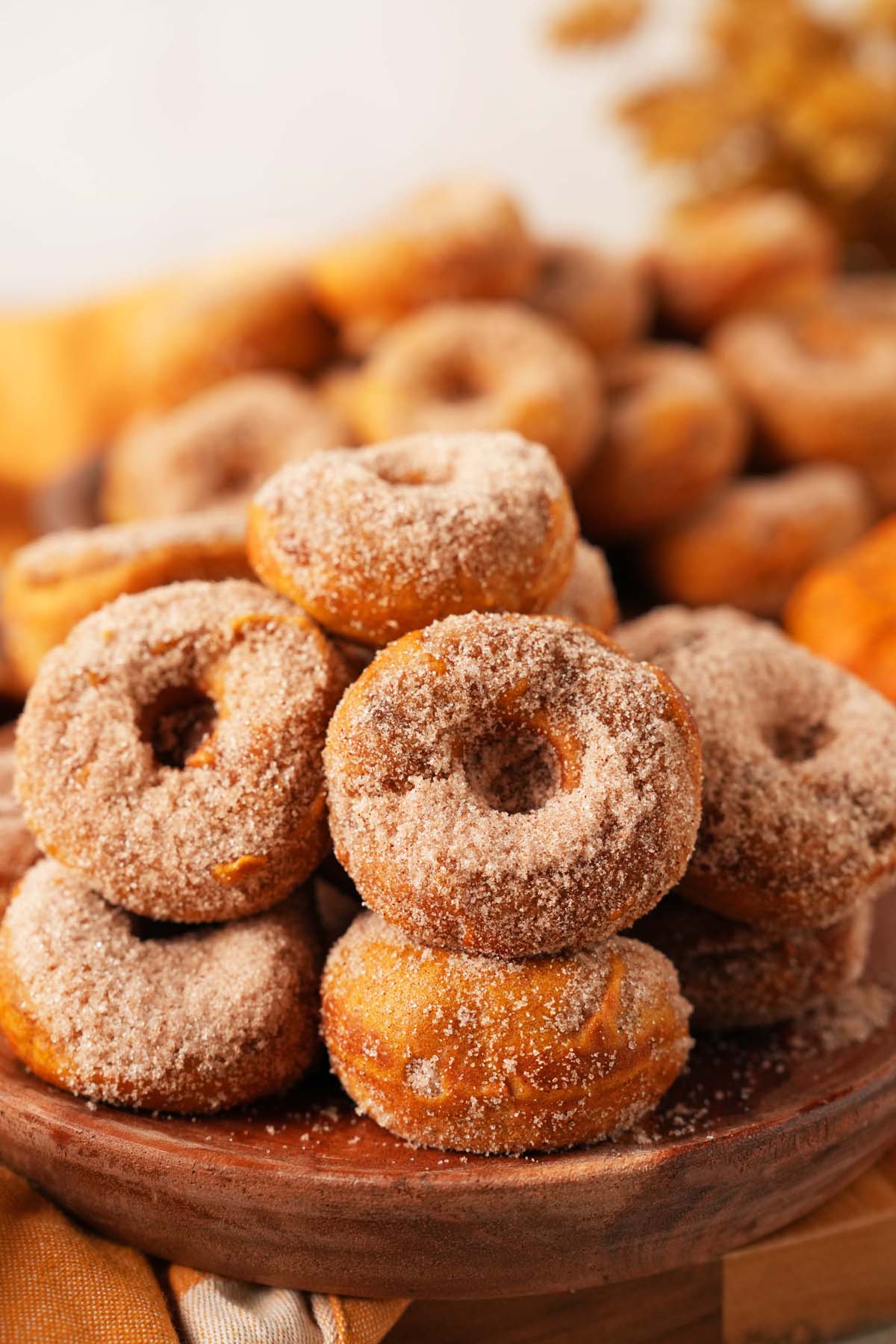 A wooden bowl stacked with pumpkin spice sugared donuts near an orange napkin and golden foliage bouquet.